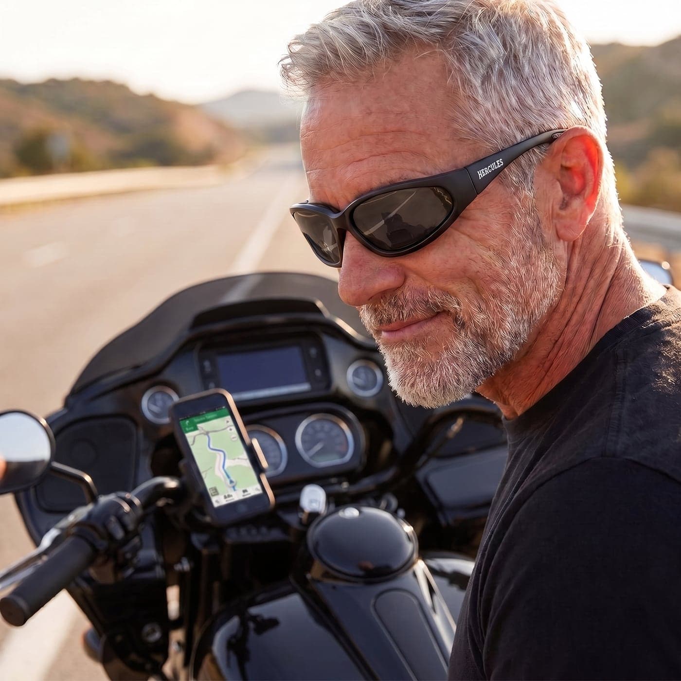 A rider stopped beside his motorcycle, wearing Hercules Rider Readers and glancing down at the map on his handlebar-mounted phone.